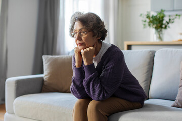 Thoughtful older woman looking in distance, thinking about future, memories, nostalgia and melancholy, feeling lonely, sitting on couch at home