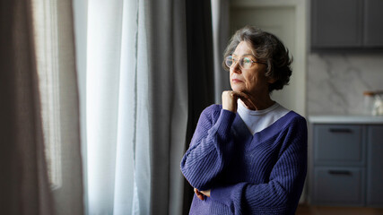 Retirement deep sad thoughts. Upset pensive senior woman standing and looking at window at home,...