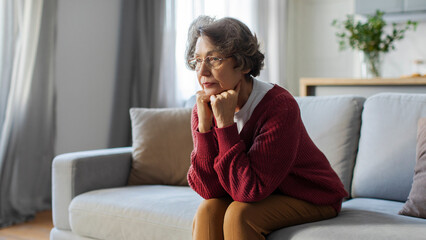 Pensive elderly lady sitting on couch at home, looking in distance and thinking lost in thoughts,...