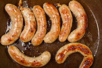 Several plump homemade sausages are sizzling in a dark frying pan, browning nicely in their own rendered fat