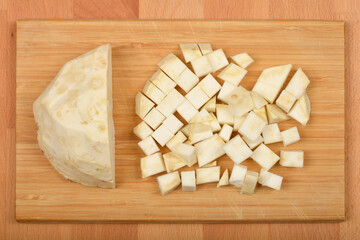 A peeled wedge and a pile of diced raw celeriac are presented on a light brown wooden cutting board, illustrating different cutting styles
