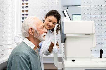 Female optician helping senior man to properly place the head on auto refractor machine to test...