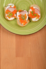 An overhead shot shows three elegant canapes with a creamy spread, vibrant orange carrot prepared as vegan salmon, radish slices, and green chives on a light green plate with a dark rim, set on a wood