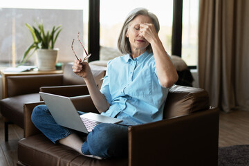 Tired senior woman taking off glasses and suffering from headache after long laptop work