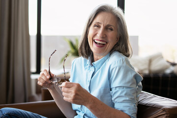 Happy senior woman laughing holding eyeglasses sitting on sofa at home