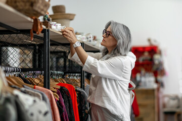 A woman with grey hair looks at vintage tableware in a boutique