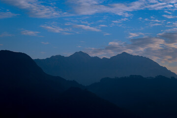 Morning scene of mountains silhouettes rest under a soft blue sky, where scattered clouds drift peacefully- capturing the calm and depth of nature's early light