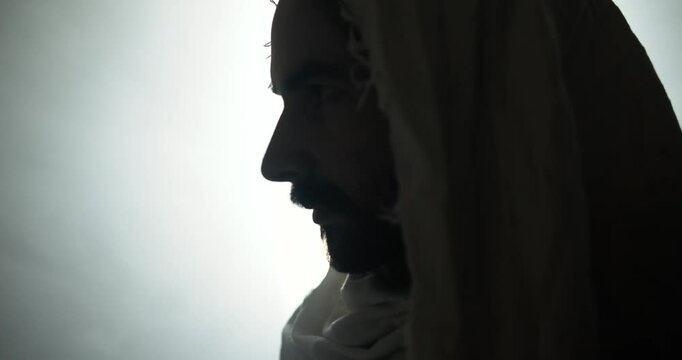 Closeup of the face of Jesus Christ resurrected on Easter Sunday inside empty tomb in bright, shining light, wearing white hood and robe. Cinematic Christian visual.