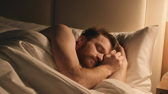 A red haired man in a hotel bed trying to sleep by the light of a side lamp.