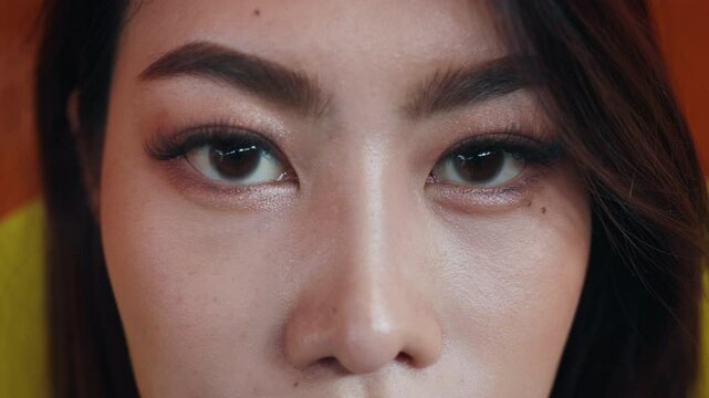 Closeup portrait of young Asian transwoman with extended eyelashes against bright rainbow flag. Pretty gender fluid female with LGBTQI community symbol looks at camera calmly