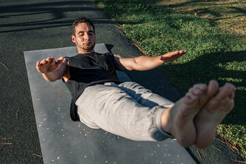 Young man performing hollow body hold on outdoor mat with arms extended and legs raised, showcasing core strength, balance, and control under sunlight