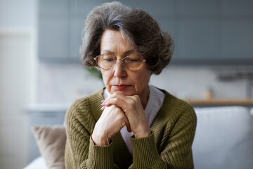 Sad lonely elder lady looking down, sitting on couch at home with chin on hands, thinking over problems, feeling sadness, apathy