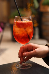 Glass with Aperol Spritz in a woman's hand. Cold refreshing alcoholic cocktail Aperol Spritz. Cocktail close-up on a blurred background. Alcoholic drink with a straw. Traditional Italian drink