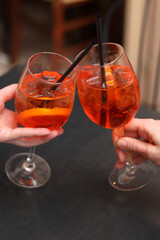 Two glasses with Aperol Spritz in the hands of girls. Cold refreshing alcoholic cocktail Aperol Spritz. Cocktails close-up with selective focus. Alcoholic drink with a straw. Traditional Italian drink