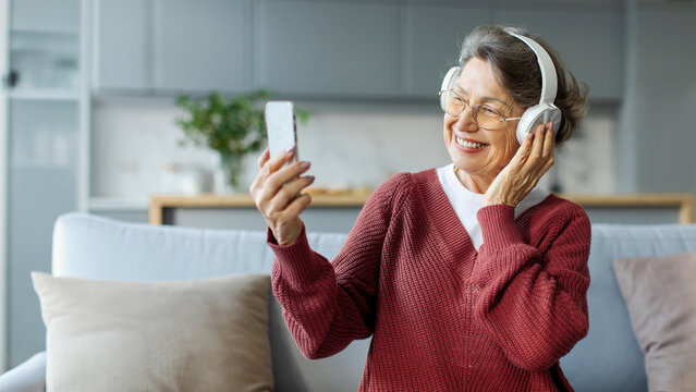 Happy senior lady in headphones sitting on couch and using smartphone, taking selfie or video calling, relaxing at free time at home, panorama, free space - Powered by Adobe