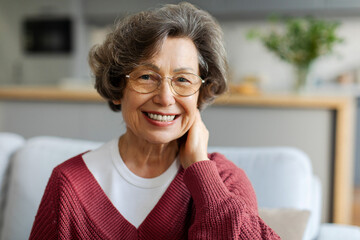 Charming senior woman with grey hair, smiling at camera while sitting on couch in living room. Beautiful carefree elderly lady spending time at home