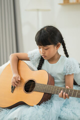 Young Girl in Light Blue Dress Playing Acoustic Guitar with Focus and Determination in Cozy Indoor...