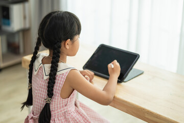 Naklejka premium Young girl engaged in digital learning on a tablet while sitting at a wooden table in a bright room, showcasing creativity and modern education tools