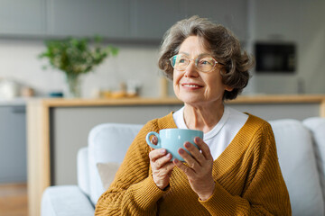 Dreamy elderly woman enjoying good morning, drinking hot coffee while resting on comfortable couch in living room, looking away and smiling