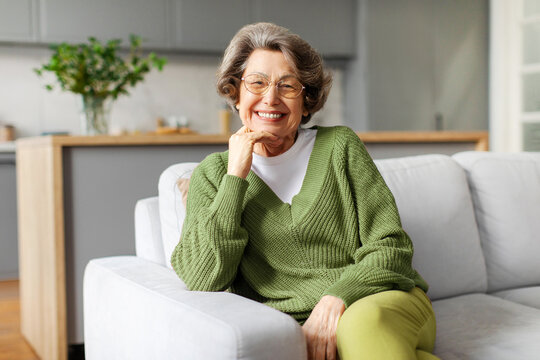 Portrait of happy senior woman relaxing on comfortable couch at home, sitting on sofa, looking and smiling at camera, enjoying retirement time