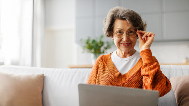 Portrait of smiling senior woman sitting on couch with laptop, looking at camera with smile, panorama with free space. Retired people technology concept
