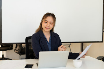 Businesswoman sitting at desk on couch in workplace or at home working on laptop and analyzing data on charts and graphs and writing on papers to make business plan and strategies for company.