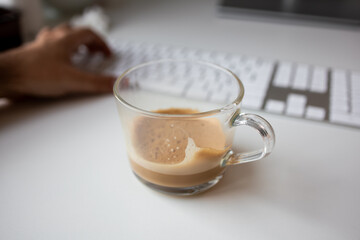Clear glass mug of coffee next to typing hands on white keyboard