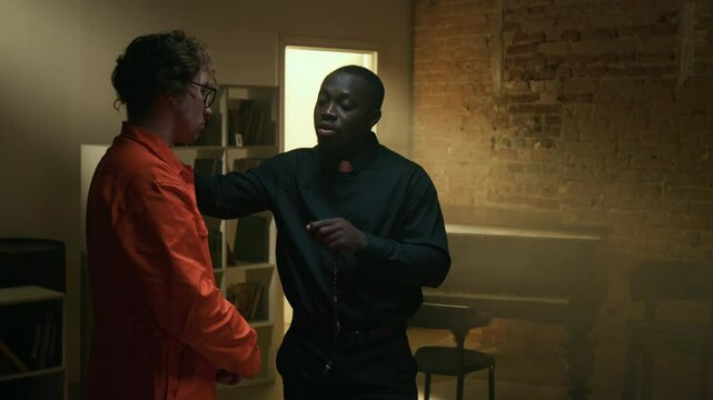 African American young pastor counselling Caucasian male convict in orange jumpsuit and holding hand on his shoulder during visit to prison