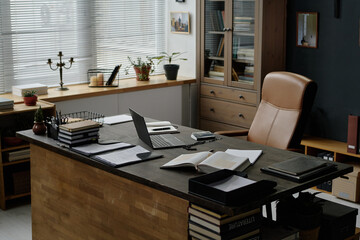 Wooden desk with black laptop, notepads, pens and opened bible on top, brown leather office chair in front of it in office of nun, copy space © AnnaStills