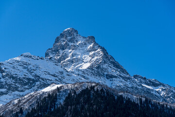 Fototapeta premium Majestic snow-capped mountain peak stands tall against clear blue sky, with rugged terrain and patches of snow covering its slopes. Karachay-Cherkessia. Dombay 2025. Nature concept for design.