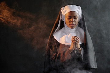 Young African American nun praying with closed eyes, she holding rosary with cross and standing in...