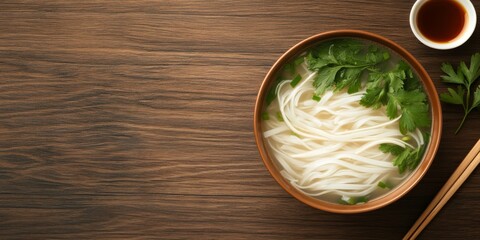 Naklejka premium Bowl of Vietnamese rice noodle soup garnished with cilantro and green onions, served with chopsticks on a rustic wood table. 