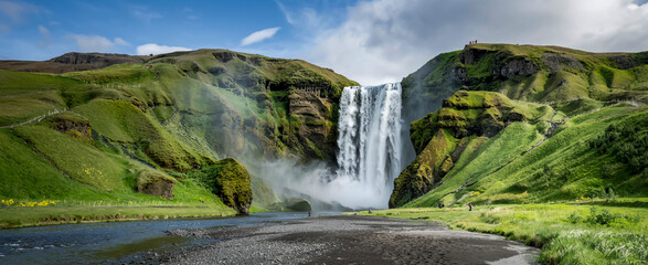 Majestic Skógafoss waterfall cascades over mossy cliffs into misty spray, framed by lush hills, volcanic black sand, vivid summer hues, and silky water flow