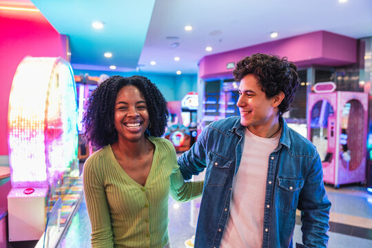 Young multi ethnic couple enjoying quality time together, laughing and playing arcade games in a vibrant, colorful entertainment center