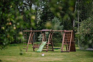 Wooden outdoor playground set with a slide, swings, and climbing frames stands on a grassy lawn surrounded by trees in a peaceful natural setting..