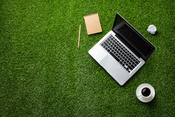 A laptop, notebook with pencil, coffee, and crumpled paper on a green grassy lawn, describing work...