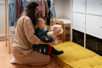Authentic cozy moment of a mother and her little daughter hugging at home, surrounded by children's clothes and toys. Real life, real emotions, real love.