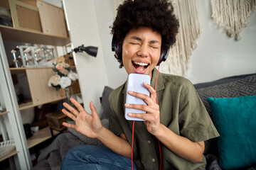 Young happy funky African American woman, funny gen z Afro girl singer in headphones singing in smartphone at home sitting on bed enjoys listening mobile music on cell phone in apps online in bedroom.