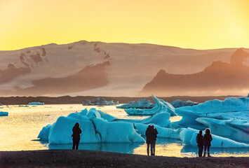 Spectators observe mesmerizing ice formations in a tranquil Ice Lagoon in Iceland during sunset. The icebergs' azure hues contrast with the warm glow of the setting sun, creating a breathtaking scene