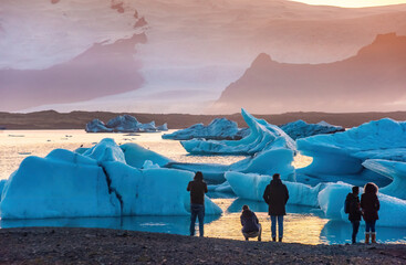 Spectators observe mesmerizing ice formations in a tranquil Ice Lagoon in Iceland during sunset. The icebergs' azure hues contrast with the warm glow of the setting sun, creating a breathtaking scene