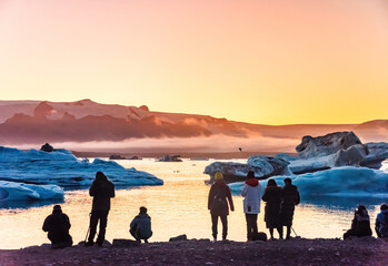 Spectators observe mesmerizing ice formations in a tranquil Ice Lagoon in Iceland during sunset. The icebergs' azure hues contrast with the warm glow of the setting sun, creating a breathtaking scene
