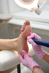 A woman on a professional pedicure procedure using an abrasive disc machine in a beauty salon. A cosmetologist in pink latex gloves cleanses the skin of the client's feet from calluses and corns