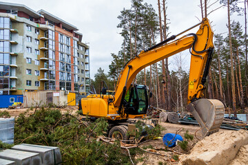 Bright yellow hydraulic excavator working on sandy ground beside a multi-story residential building under construction, with fallen tree branches and forest in background