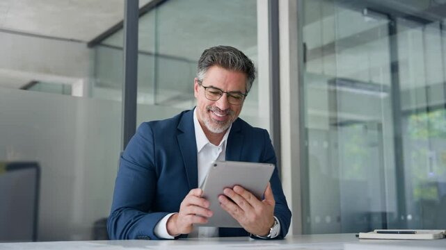 Positive senior businessman entrepreneur sitting at desk working on financial sales project, looking smiling at screen. Mature 50s Latin business man ceo manager using tablet pad computer in office