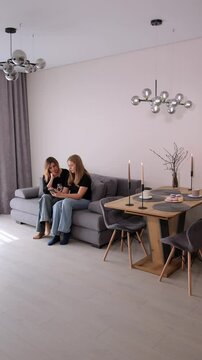 Vertical video. Two adult women friends sitting on a sofa in the living room and looking at a smartphone. New minimalist interior of a studio kitchen and a table with tea and macaroons.