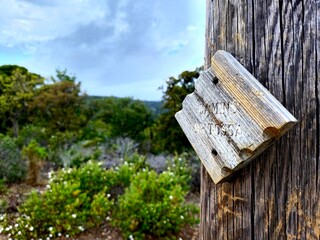 Old, weathered wooden hiking sign nailed to a post, indicating the direction to sertosa, amidst a backdrop of lush green vegetation and a cloudy sky
