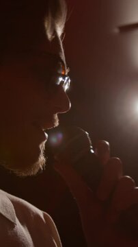Vertical close-up shot of young man with beard and glasses talking into microphone on dimly lit stage