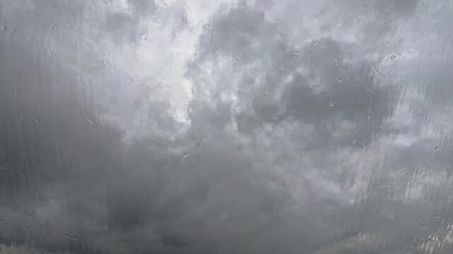 Nahaufnahme von Regentropfen, die an einer Fensterscheibe runterlaufen, mit dramatisch dunklen Regenwolken im Hintergrund. Tr&uuml;be Wetterstimmung, Zeitraffer, Metrologie, Melancholie, Herbst, Natur
