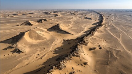 Aerial view of a desert landscape with a long, winding rock formation stretching across the sand dunes under bright sunlight.