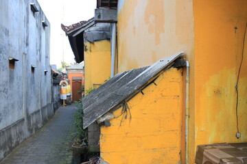 Yellow building with a roof and a brick wall. The building is in a narrow alleyway